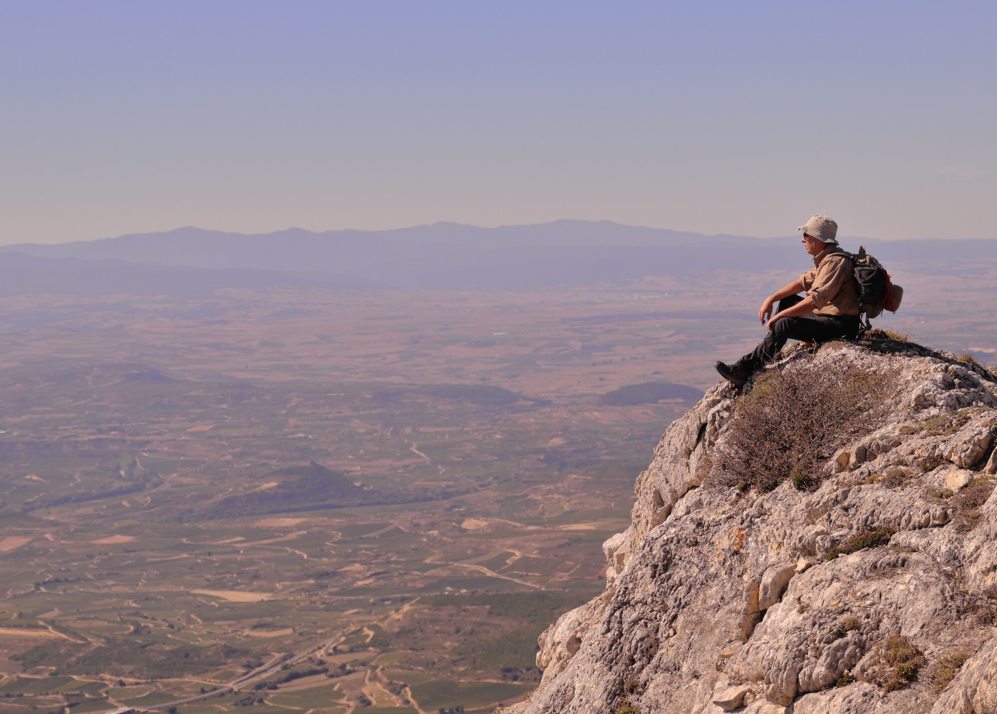 This image shows a mountaineer resting on a rocky peak and taking in the view across a wide valley.